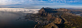 Luftaufnahme einer Küstenstadt zwischen dunkelblauem Ozean und zerklüfteten Bergen, teilweise in Nebelschwaden gehüllt, Kapstadt, Westkap, Südafrika.