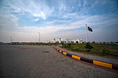 View of the Kartarpur Corridor gleams white under a vast sky, a Pakistani flag fluttering proudly, bordered by a meticulously paved road, Kartarpur, Punjab, Pakistan.