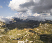 Luftaufnahme der zerklüfteten Landschaft von Campo Imperatore, eingebettet unter dramatischen Wolken, mit kurvenreichen Straßen und einem Spiel aus Licht und Schatten, Gran Sasso, Abruzzen, Italien.