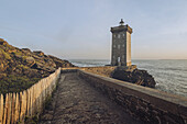 View of the Kermorvan lighthouse standing proudly against the Atlantic, a stone path leading to it framed by weathered rocks and a rustic wooden fence, Le Conquet, Bretagne, France.