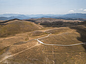 Luftaufnahme von sonnenverbrannten Hügeln, die sich auf ferne Berge zubewegen und von einer hellen Straße unter einem weiten Himmel halbiert werden, Gran Sasso, Abruzzen, Italien.