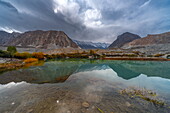 View of serene lake reflecting rugged mountains under a dramatic sky, showcasing nature's raw beauty and contrasting textures, Hunza Nagar, Gilgit Baltistan, Pakistan.