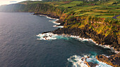 Luftaufnahme der dramatischen Küstenlinie, wo grüne Klippen auf den stürmischen Ozean treffen. Die dunklen vulkanischen Felsen kontrastieren mit dem weißen Schaum der anbrandenden Wellen, Azoren, Portugal.