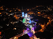 Aerial view of a medieval town square glows with vibrant Christmas lights illuminating the church, roofs, and festive decorations, Turckheim, Grand Est, France.