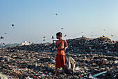 Chattogram, Bangladesh - 03 November 2019: View of a boy standing amidst a vast landfill, holding a frame, with birds circling above, against a clear sky.