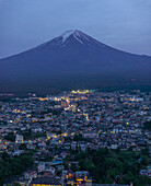 Aerial view of Mount Fuji looming majestically over a town, lights twinkling against the dusk, a serene symphony of nature and urban life, Fujiyoshida, Yamanashi, Japan.