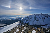 View of sun shining brightly over snow-covered mountain peaks and valleys under a blue sky, creating a serene and majestic winter scene, Vysoké Tatry, Prešov Region, Slovakia.