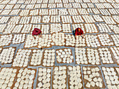 Bogura, Bangladesh - 04 May 2023: Aerial view of women in red garments amidst a textured landscape of drying food, forming an intricate pattern under the sun.