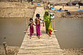 Bogura, Bangladesch - 02. Juni 2024: Blick auf eine Frau und Kinder, die eine Bambusbrücke überqueren. Ihre farbenfrohe Kleidung steht im Kontrast zu den erdigen Tönen des Flusses und der sandigen Ufer.