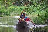 Bogura, Bangladesch - 28. Juli 2017: Blick auf ein Boot, das durch das Wasser gleitet und von einem Mann gesteuert wird, der Kinder inmitten von üppigem Grün trägt.