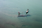 Rangpur, Bangladesh - 30 November 2019: Aerial view of two figures standing near a submerged structure, casting long shadows on the tranquil, pale green water as one navigates a small wooden boat.
