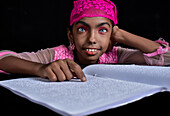 Mirsharai, Bangladesh - 20 September 2020: View of a young girl with striking blue eyes, adorned with a vibrant pink headscarf, intently reading a braille book against a stark black backdrop.