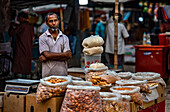 Bogura, Bangladesh - 21 March 2022: View of a vendor at a bustling market, his weathered face framed by sacks of nuts and grains, under soft light.