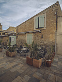 View of a rustic stone building with light green shutters, a charming outdoor cafe with potted plants under a cloudy sky, Gruissan, Occitanie, France.