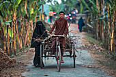 Bogura, Bangladesh - 25 December 2020: View of a young boy cycling a wooden cart laden with firewood, accompanied by a woman in a black niqab along a rural road.