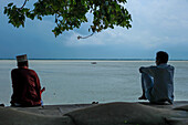 Chandpur, Bangladesh - 28 June 2019: View of two men contemplating life along the Meghna River, their silhouettes framed by the cool, overcast sky and rippling water.