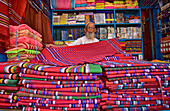 Bogura, Bangladesh - 11 March 2018: View of vibrant stacks of colorful textiles and a shopkeeper with a white beard displaying his wares in a market stall.