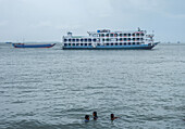 Chandpur, Bangladesch - 28. Juni 2019: Blick auf eine große Fähre und ein kleineres Boot, die durch das trübe Wasser fahren, während Kinder in Ufernähe schwimmen.