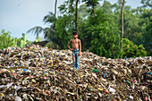 Bogura, Bangladesh - 18 August 2020: View of a young boy standing atop a vast, colorful mountain of discarded waste, contrasting sharply with the lush green foliage behind him.