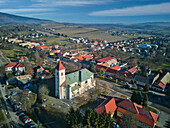 Luftaufnahme einer Kirche mit grünen Dächern und einem Turm mit roter Spitze inmitten eines Dorfes in einem Tal, dessen Gebäude lange Schatten werfen, Detva, Region Bansk? Bystrica, Slowakei.