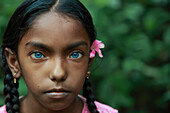 Mirsharai, Bangladesch - 20. September 2020: Blick auf ein junges Mädchen mit bezaubernden blauen Augen und geflochtenem Haar, das mit einer zartrosa Blume geschmückt ist, vor einem Hintergrund aus üppigem Grün.