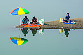 Bogura, Bangladesh - 16 December 2017: View of men fishing along the serene riverbank, their vibrant umbrellas mirrored in the tranquil water.