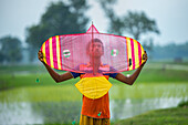 Bogura, Bangladesh - 11 July 2020: View of a young boy holding a brightly colored kite against the soft green backdrop of rural fields.