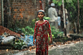 Bogura, Bangladesh - 06 October 2020: View of a young girl braving the downpour on a street, her red dress a vibrant splash against the muted tones of the rain-soaked landscape.