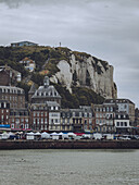 Blick auf die Gebäude der Stadt mit ihren braunen und grauen Fassaden am Fuße der hoch aufragenden weißen Klippen unter einem bedeckten Himmel, Le Tréport, Normandie, Frankreich.