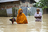 Satkania, Bangladesh - 09 August 2023: View of a woman in an orange shawl wading through floodwaters, clutching belongings, with an elderly man standing nearby amidst submerged homes.
