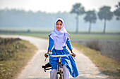 Bogura, Bangladesh - 09 February 2020: View of a young girl in a blue school uniform and white hijab, cycling along a sun-drenched road framed by lush greenery and distant palms.