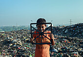 Chattogram, Bangladesh - 03 November 2019: View of a young boy standing amidst a vast, sprawling landfill, his gaze piercing through a black plastic frame.