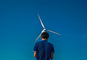 Feni, Bangladesh - 27 May 2019: View of a lone figure silhouetted against a vibrant blue sky, gazing up at the wind turbine's blades in motion.