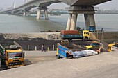 Dhaka, Bangladesh - 17 May 2019: View of weary laborers resting beside vibrant trucks near the Buriganga River, under the shadow of a colossal bridge.