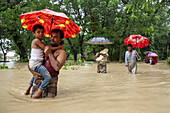 Satkania, Bangladesch - 09. August 2023: Blick auf einen Vater, der sein Kind durch die Fluten trägt. Der leuchtend rote Regenschirm steht in starkem Kontrast zu den trüben Fluten.