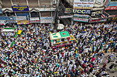 Chattogram, Bangladesh - 08 October 2022: Aerial view of a dense crowd gathered around a decorated vehicle, banners waving amidst the urban sprawl, a sea of faces under a web of overhead cables.
