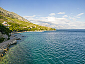 Luftaufnahme des türkisfarbenen Wassers der Adria, das sanft die felsige Küste unter den zerklüfteten, sonnenverwöhnten Bergen von Artina Beach, Lokva Rogoznica, Kroatien, küsst.