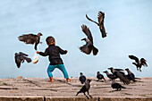 Bogura, Bangladesh - 20 November 2021: View of a child in vibrant blue trousers scattering food for a flock of crows against the pale sky on a stone embankment.