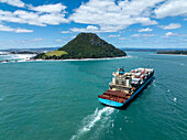 Tauranga, Neuseeland - 16. Januar 2022: Aus der Vogelperspektive sieht man ein riesiges Containerschiff, das sich unter strahlendem Himmel durch das türkisfarbene Wasser auf den Mount Maunganui zubewegt.