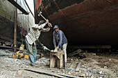 Dhaka, Bangladesh - 05 November 2021: View of two men working on metal with a hammer and anvil against the backdrop of massive, rusted ships, a scene of raw labor and industrial grit.