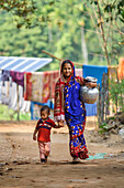 Bogura, Bangladesch - 10. Dezember 2019: Blick auf eine Mutter in einem leuchtend blauen Sari und ihr Kind, die einen staubigen Weg entlanggehen, einen silbernen Topf auf ihrer Hüfte balancierend, inmitten des ländlichen Charmes von Bogura.