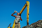 Chattogram, Bangladesh - 03 November 2019: View of a boy drinking water against a backdrop of a colossal yellow excavator and a mountain of trash.