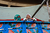 Chattogram, Bangladesh - 21 November 2018: View of two young girls with vibrant headscarves peek over the ornate, blue metal truck bed, their expressions a mix of curiosity and shyness.