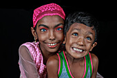 Mirsharai, Bangladesch - 20. September 2020: Blick auf zwei strahlend lächelnde Kinder, deren auffallend blaue Augen sich von ihrer dunklen Haut und dem tiefschwarzen Hintergrund abheben.