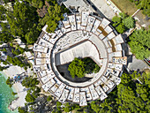 Aerial view of a circular, decaying structure surrounded by lush greenery and turquoise waters, a stark contrast of nature reclaiming man's creation, Krvavica, Split-Dalmatia County, Croatia.