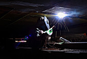 Dhaka, Bangladesh - 25 October 2019: View of a worker welding beneath a structure, bright sparks contrasting with the dark shadows.