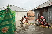 Kurigram, Bangladesch - 21. Juni 2022: Blick auf die Überschwemmungen, in denen sich Wellblechhäuser spiegeln, während ein Kind Wasser pumpt und eine Frau in dem überschwemmten Dorf Wasser sammelt.