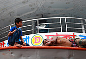 Dhaka, Bangladesh - 17 May 2019: View of a young boy sitting, gazing pensively as a man rests shirtless on the deck of a vessel, under the boat's ornate ceiling.