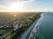 Aerial view of sun-kissed sands meet the rhythmic waves of the ocean, contrasting with the lush greenery and the distant mountain, Papamoa, Bay of Plenty Region, New Zealand.