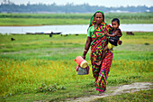 Bogura, Bangladesch - 07. Mai 2017: Blick auf eine Mutter, die ihr Kind und Behälter über einen grasbewachsenen Weg in der Nähe eines ruhigen Flussufers trägt.